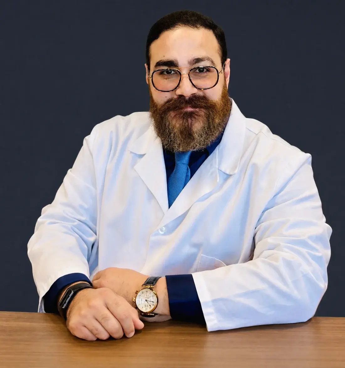 Jules Bakhos, host of PrevPod, in a white coat with his arms folded on a wooden table, against a dark blue wall